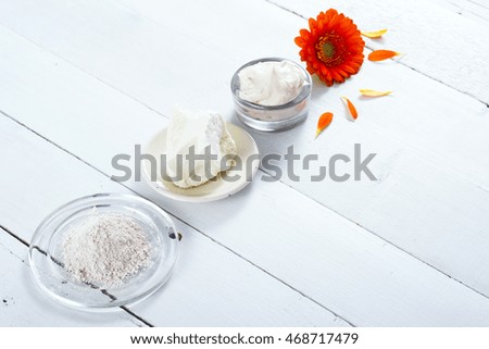 sea clay powder, moisturizer and raw shea butter block with gerbera flower on white wooden table background
