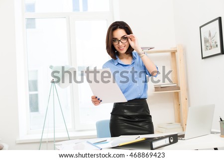 Brunette girl in blue shirt and black skirt is standing near table in office. She holds glasses  on face and paper in hand. She is looking to the camera.