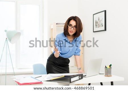 Brunette girl in blue shirt and black skirt is standing near table in office. She rested her hand on the table. She is looking to the camera very seriously.