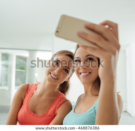 Cute girls sitting at desk and taking self portraits using a smart phone, they are smiling at camera and posing