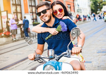 Young beautiful happy couple rides around town on a vintage scooter, a romantic date, fashionable casual wear, sunglasses, girl holding a bouquet of flowers and embraces the guy, laughing, urban style