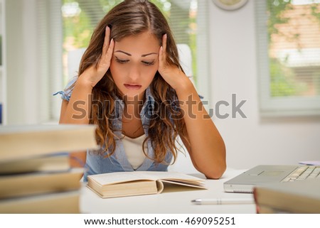 Beautiful young woman learning in the library. She is reading book and holding her head.