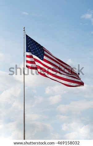 A single American flag flying high as the wind helps it to spread its colors in the late afternoon sun. Fluffy clouds and a blue sky make the perfect background. Vertical format with copy space.
