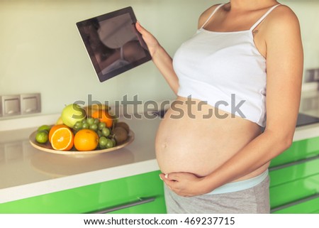 Cropped image of beautiful pregnant woman holding a digital tablet and keeping a hand on her bare tummy while standing in the kitchen at home