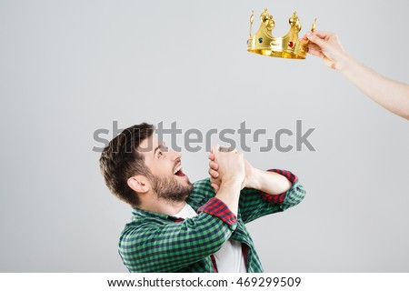 Man in green checked T-shirt and crown above his head. Looking at crown, very surprised. Waist up, profile, hands together. Indoors, studio