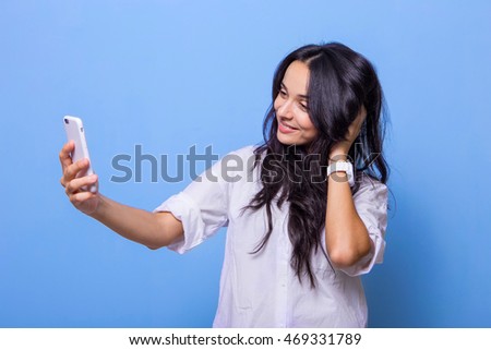 
Portrait of a beautiful young smiling girl who is holding a smartphone and does with it selfie on a blue background .