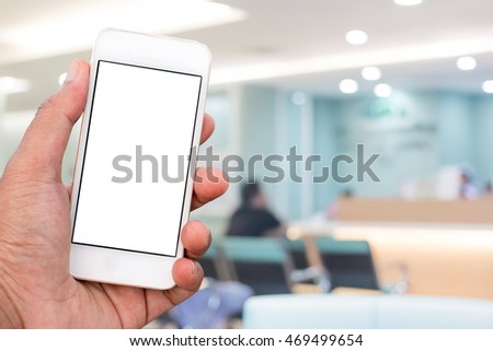 Hand holding mobile smart phone with blank screen in vertical position, blurred people in hospital background - mockup template