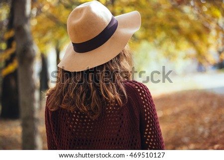 Rear view of brunette girl in autumn/fall park in brown hat, sweater and trousers. Back view of autumn portrait of woman outdoors with curly hair Royalty-Free Stock Photo #469510127