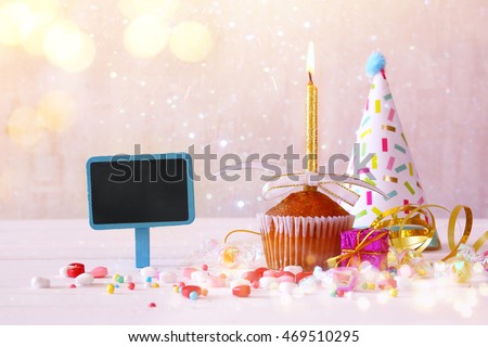 Birthday concept with cupcake and candle next to little chalkboard on wooden table. Glitter overlay. Selective focus