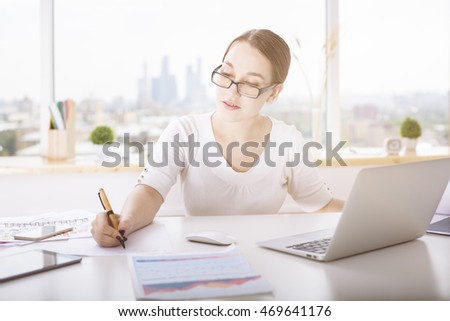 Portrait of attractive young lady doing paperwork at office desk with laptop, tablet, business report and other items