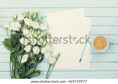 Morning coffee cup, empty paper list, pencil, and bouquet of white flowers eustoma on blue rustic table from above. Woman working desk. Cozy breakfast. Flat lay styling.