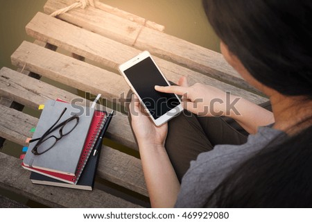 Weekend morning lifestyle.Young woman touching on mobile phone screen while sitting outdoor beside river with notebooks, pen, and glasses in morning time.Freelance working and phone addiction concept