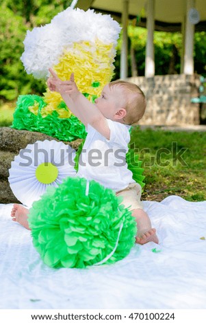 Happy little boy celebrating his first birthday sitting on a rug in the park below a colorful number 1 decoration in white, green and yellow