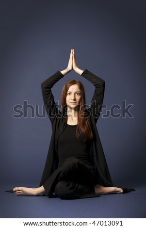 Young lady practicing yoga in Siddhasana posture with eyes opened in black clothes on blue background.