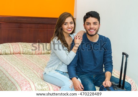 Young charming hispanic couple wearing casual clothes sitting on bed next to suitcase, hostel guest concept