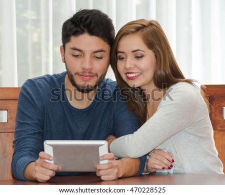 Young charming couple seated by table watching tablet screen while embracing, bot happy and smiling, hostel concept
