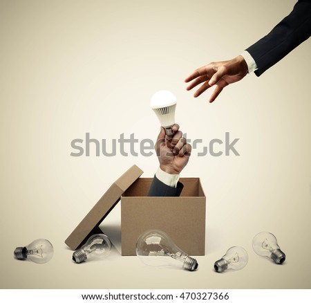 Hand of a businessman holding a turned on LED light bulb coming out from a brown paper box surrounded by old incandescent light bulbs / Business with new idea and innovation concept