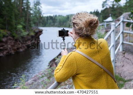 Young woman in a yellow coat photographing river on a smartphone with selfie stick
