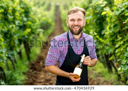 Portrait of handsome wine maker in apron with bottle of white wine standing on the vineyard Royalty-Free Stock Photo #470436929