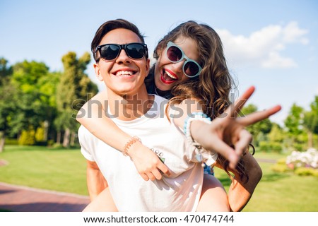 Portrait of cute couple having fun in park. Pretty girl with long curly hair is riding on back of handsome guy. They wear sunglasses and smile to camera.