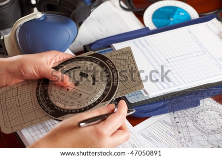 Pilot equipment with airplane pilot hand using flight computer for navigation calculations, other tools like protractor, kneepad with charts and professional headset