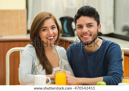 Young charming couple seated by breakfast table smiling to camera, fruits, juice and coffee placed in front, hostel environment