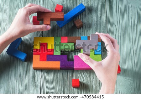 Hand holding wooden puzzle element. Hand sets the last element of the puzzle. The concept of logical thinking. Geometric shapes on a wooden background. Tetris toy wooden blocks. Royalty-Free Stock Photo #470584415