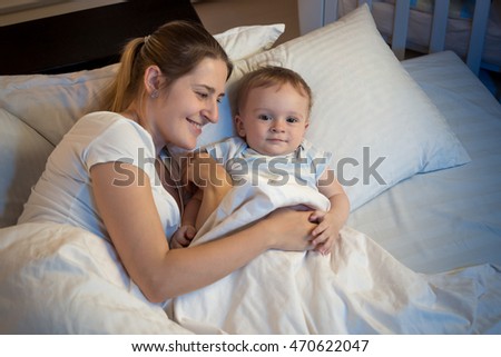 Portrait of beautiful smiling woman lying in bed at night and hugging her baby boy