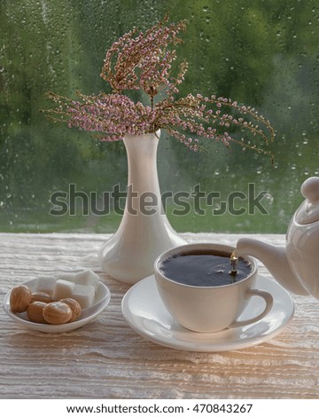 English black tea in white bowl. Tea is poured from the kettle. On the table, caramel sugar and a bouquet of Heather in a vase. Outside, the rain. Rain drops on glass.