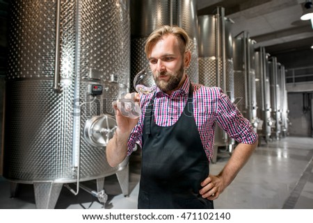 Handsome wine maker in working apron checking wine quality at the manufacturing with metal tanks for wine fermentation. Wine production at the modern manufacture Royalty-Free Stock Photo #471102140