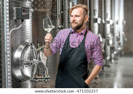 Portrait of a handsome wine maker in working apron with the wine glass at the manufacture with metal tanks for wine fermentation. Wine production at the modern factory Royalty-Free Stock Photo #471102257