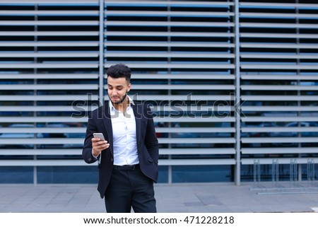 Entrepreneur uses phone mobile smart looking at the camera. Young handsome businessman arab muslims in business center wearing dressed in black elegant suit on building background.