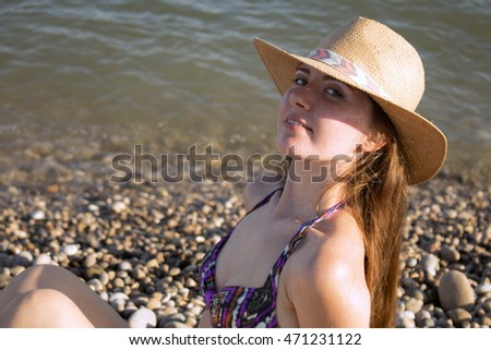 Beautiful young woman with sun hat sitting and smiling on the beach. Sea in background. horizontal