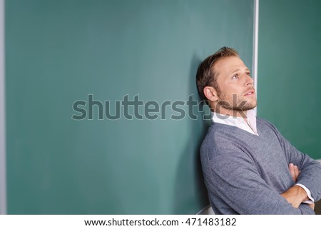 Young male teacher or post graduate university student standing daydreaming leaning back with folded arms against a clean blackboard with copy space staring into the air