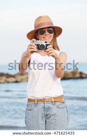 Portrait of a middle age woman wearing straw hat and standing on the beach while taking a photo with her vintage camera. 