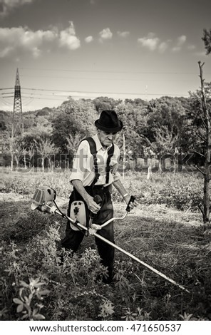 Old farmer using a petrol brush cutter to mow long grass and flowers. Black and white picture.