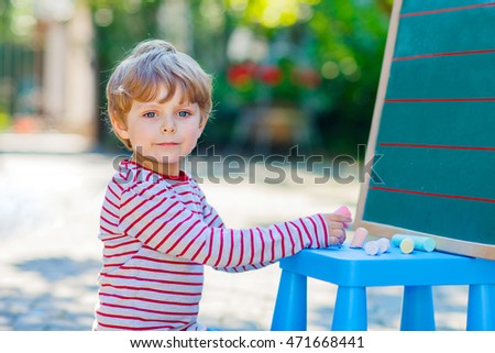 Adorable funny little kid boy at blackboard practicing writing letters, outdoor school or nursery. Child having fun with learning. Back to school concept.