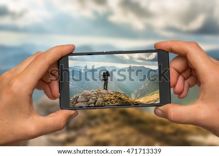 Woman hands with mobile cell phone to take a photo of young man standing on a rock and looking at a beautiful mountains 