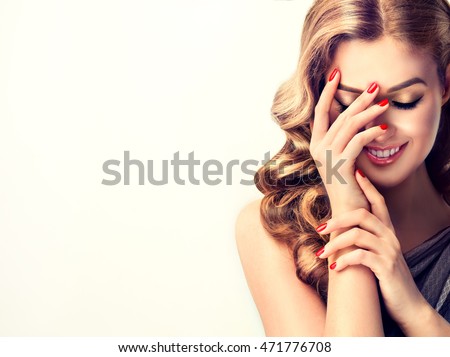 Beautiful woman with curly hair and red nails manicure . Girl laughs shyly closes her face with a hand .
