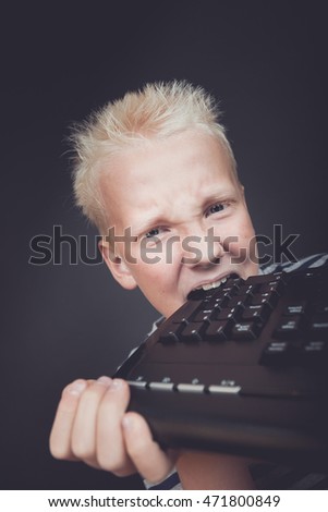 Angry boy with blond hair holding keyboard bites into it against a dark background