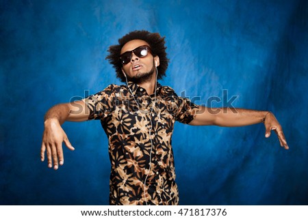 African man listening music in headphones, dancing over blue background.