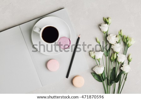 Morning cup of coffee, empty notebook, pencil, white flowers and cake macaron on gray table from above. Beautiful breakfast. Flat lay.