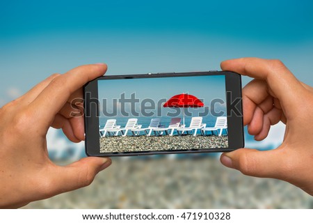 Woman hands with mobile cell phone to take a photo of sunbathing plastic beds and red umbrella on the beach
