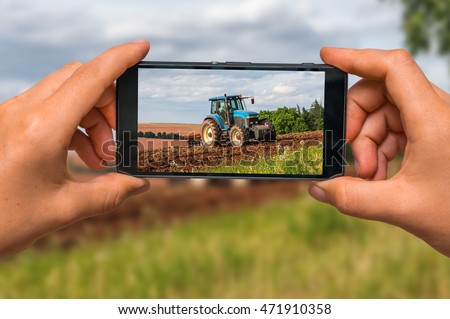 Woman hands with mobile cell phone to take a photo of tractor at work on a field