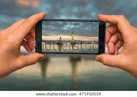 Woman hands with mobile cell phone to take a photo of evening seascape with lighthouse in harbor