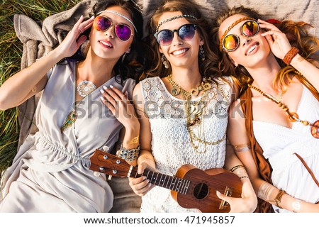 Three cute hippie girl lying on the plaid outdoors, best friends having fun and laughing, play ukulele, sunglasses, feathers in their hair, bracelets, flash tattoo, indie, Bohemia, boho style top view Royalty-Free Stock Photo #471945857