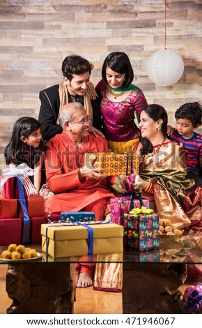 3 generations of Indian family celebrating Diwali festival or birthday by exchanging gifts and sweets while sitting on Sofa, Indoors
