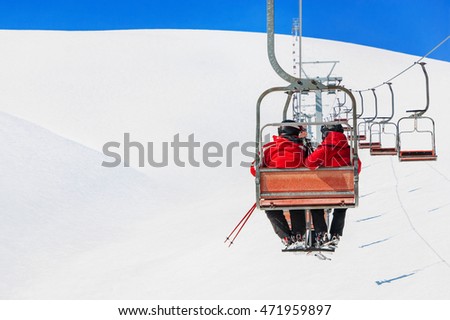 Skiers couple in red skisuits go on a ski lift at chairlift ropeway against snowy mountains landscape of ski slopes and blue sky - winter holidays concept