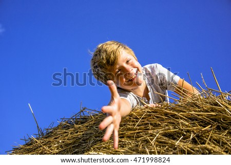 Smiling boy on a haystack on the background of blue sky pulls the arm to the camera
