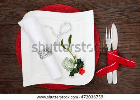 Christmas dinner table setting with white square porcelain plate, napkin, silver bead strand, holly, mistletoe and cutlery with red ribbon over oak wood background.